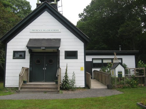A white school building with a black roof, featuring a sign that reads "Prudence Island School" and a wooden walkway.