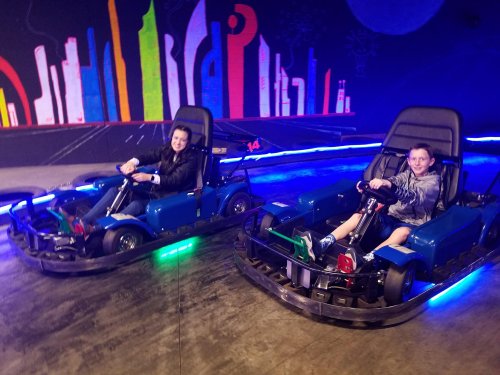 Two children in go-karts, smiling and racing on a colorful indoor track with neon lights.