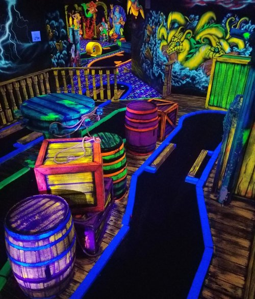 A vibrant, neon-lit mini-golf course featuring colorful barrels, crates, and whimsical decorations.