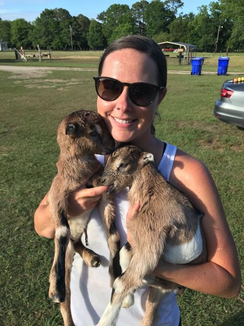 A woman in sunglasses holds two baby goats, smiling in a grassy outdoor setting.