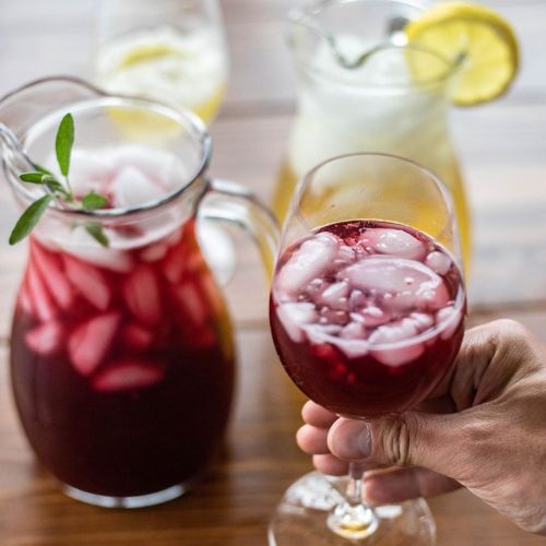A hand holds a glass of red drink with ice, surrounded by pitchers of various beverages on a wooden table.