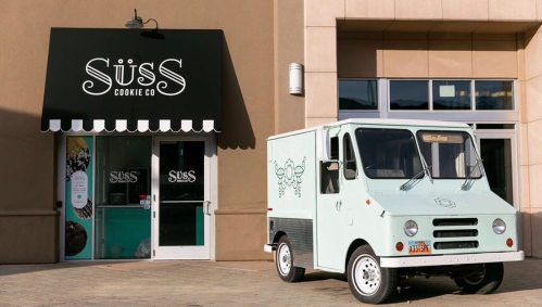 A light blue cookie truck parked outside the Süß Cookie Co. storefront with a black awning.