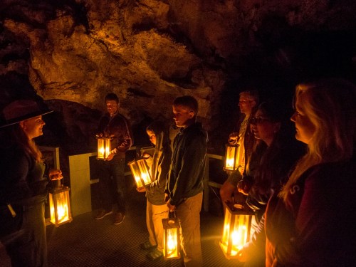 A group of people holding lanterns in a dark cave, listening to a guide.