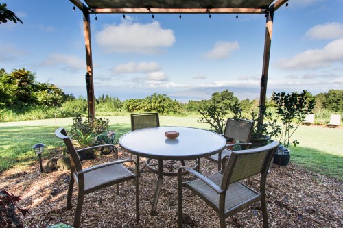 A patio with a round table and chairs overlooks a lush garden and distant ocean under a blue sky with clouds.