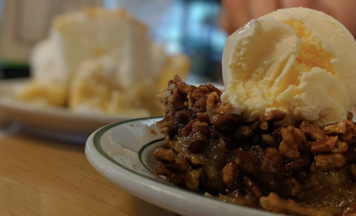 A close-up of a slice of pecan pie topped with a scoop of vanilla ice cream on a plate.