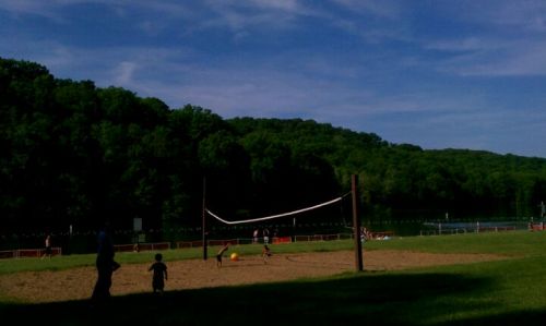 A sandy volleyball court by a lake, with children playing and green trees in the background under a blue sky.
