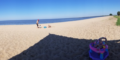 A sandy beach with a calm sea, featuring two children playing and an adult nearby under a clear blue sky.