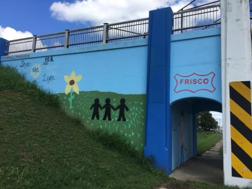 Colorful mural under a bridge featuring silhouettes of children holding hands, a flower, and the word "FRISCO."