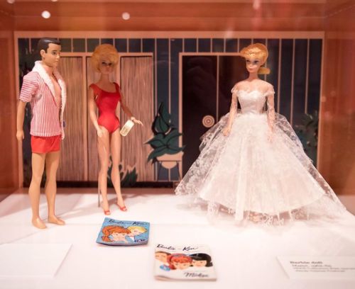 Two vintage dolls, a Ken in swimwear and a Barbie in a wedding dress, displayed with magazines in a showcase.