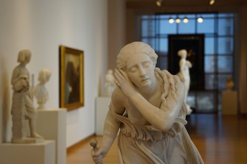 A marble sculpture of a woman with a serene expression, set in an art gallery with other sculptures in the background.