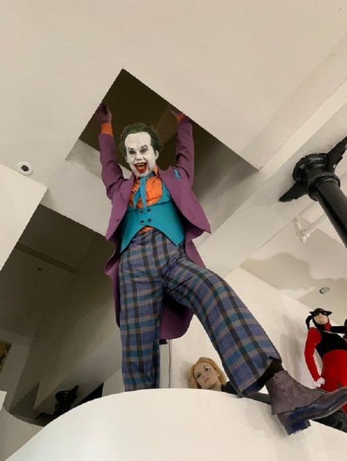 A costumed figure resembling a clown hangs from a ceiling opening, wearing a purple suit and colorful attire.