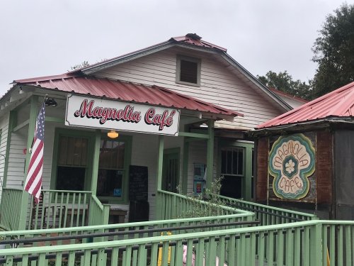 A charming café with a red roof, green porch, and signs reading "Magnolia Café" in a quaint setting.