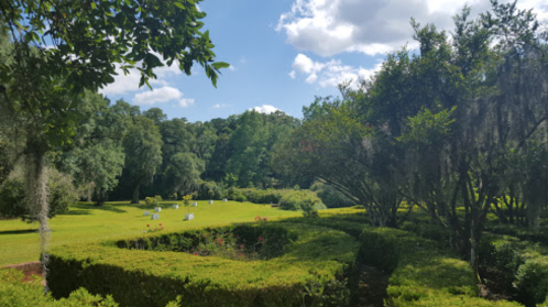 Lush green garden with neatly trimmed hedges, trees, and a bright blue sky dotted with clouds.