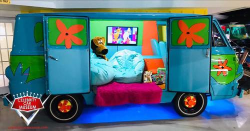 A colorful van with a cozy interior, featuring blue cushions and a TV, displayed at a car museum.