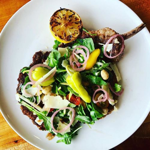 A plate featuring a breaded chop topped with a fresh salad, pickled onions, and a grilled lemon half.