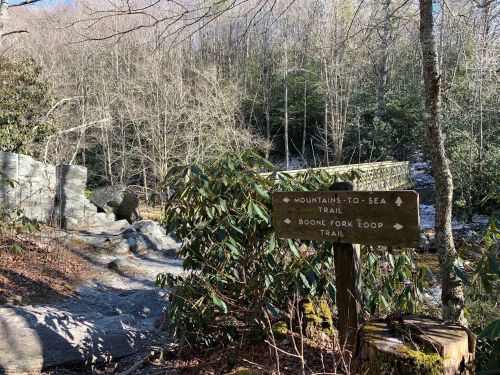 A wooden signpost at a trail intersection in a forest, indicating the Mountains-to-Sea Trail and Boone Fork Loop Trail.