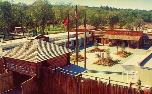 A vintage view of a themed outdoor area featuring wooden structures, flags, and landscaped gardens.