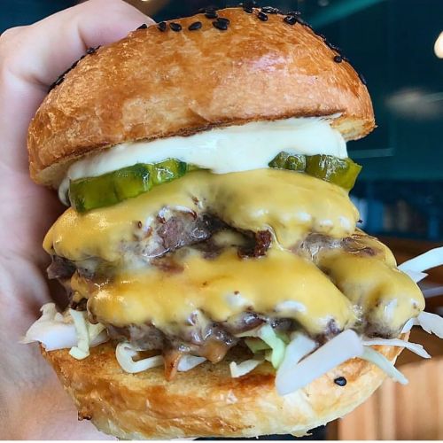 A hand holding a stacked cheeseburger with melted cheese, pickles, and mayonnaise on a sesame seed bun.
