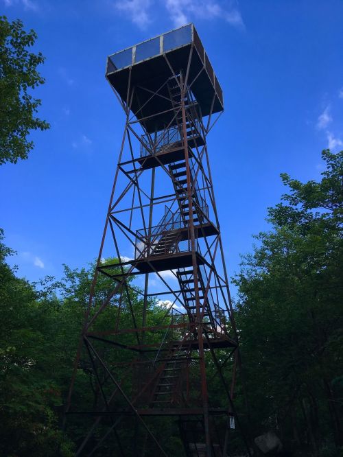 A tall, rusted observation tower surrounded by green trees under a blue sky with scattered clouds.