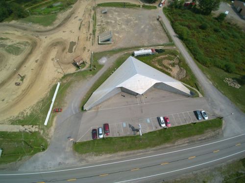 Aerial view of a modern, triangular building with a parking lot, surrounded by dirt tracks and greenery.