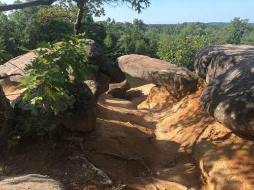 Rocky landscape with large boulders and a sandy path leading to a serene, wooded area in the background.