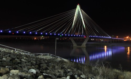 A illuminated cable-stayed bridge at night, reflecting in the water below, with rocks and grass in the foreground.
