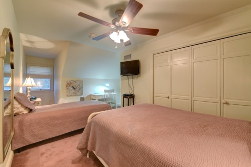 Cozy bedroom with two twin beds, a ceiling fan, and a TV, featuring light-colored walls and a small desk.
