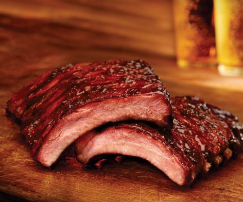 Close-up of tender, glazed barbecue ribs on a wooden surface, with a blurred background of beer glasses.