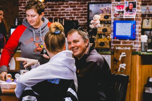 A group of people chatting at a cozy café, with one person smiling and another serving food. Warm, inviting atmosphere.