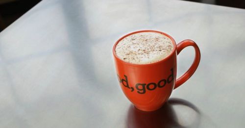 A bright orange mug with the words "good, good" filled with frothy coffee sits on a reflective surface.
