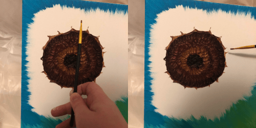 A hand holds a paintbrush near a detailed sunflower painting on a canvas with blue and green background.