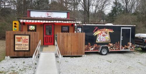 A food truck and small building with a sign reading "Open" and "Luv 4 Shak," surrounded by trees and gravel.
