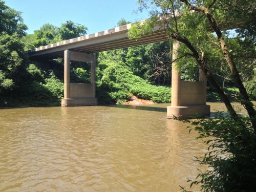 A concrete bridge spans a river, surrounded by lush greenery and trees on a sunny day.
