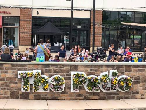 A lively outdoor event at "The Patio," with a crowd enjoying a performance surrounded by flower-adorned signage.