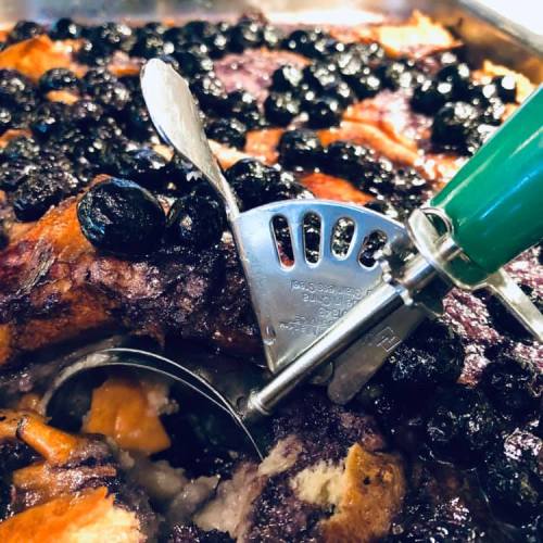 A close-up of a baked dish topped with blueberries, featuring a metal serving tool with a green handle.