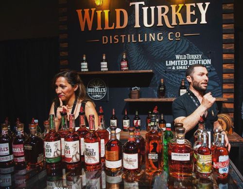 A bar display featuring various Wild Turkey whiskey bottles, with two bartenders preparing drinks.