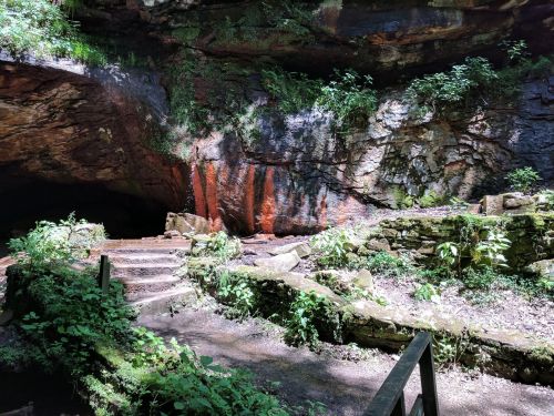 A serene cave entrance surrounded by lush greenery and rocky formations, with steps leading into the cave.
