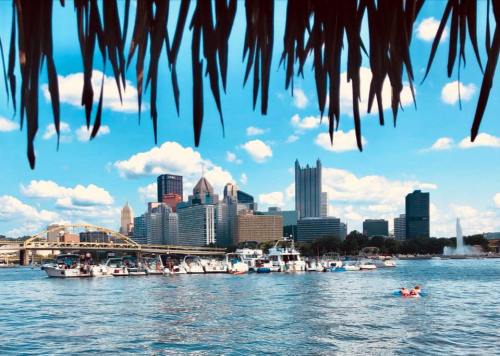 View of Pittsburgh skyline from a waterfront, with boats on the river and a clear blue sky dotted with clouds.