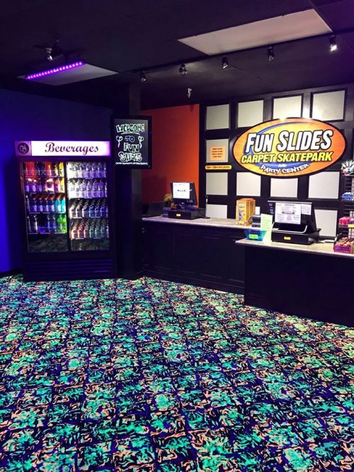 Interior of a vibrant carpet skatepark with a beverage fridge, snack counter, and colorful flooring under blacklight.