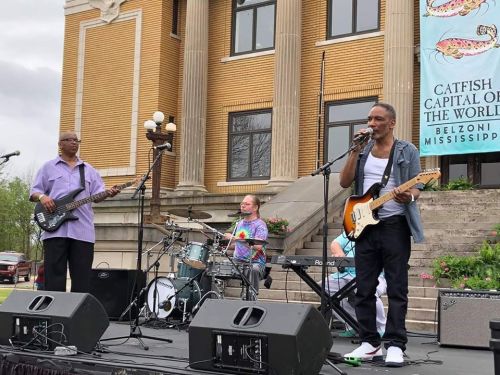 A band performs on stage outdoors, featuring a guitarist, bassist, drummer, and keyboardist in front of a historic building.