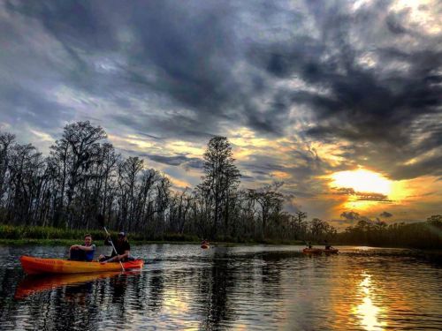 Two kayakers paddle on a calm river at sunset, surrounded by trees and dramatic clouds reflecting on the water.