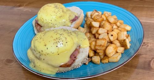 A blue plate with two eggs Benedict on English muffins and a side of seasoned potato cubes.