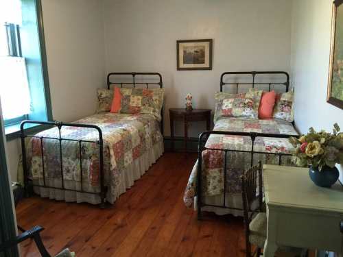 A cozy bedroom with two metal-framed beds, floral quilts, a small table, and a vase of flowers.