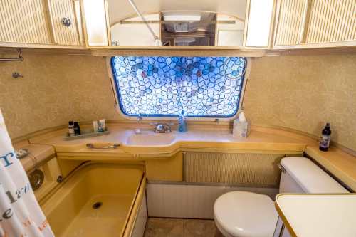A vintage bathroom with a yellow bathtub, sink, and toilet, featuring a patterned window curtain and various toiletries.