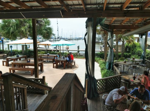 Outdoor dining area at a marina, featuring wooden tables, umbrellas, and views of boats in the water.