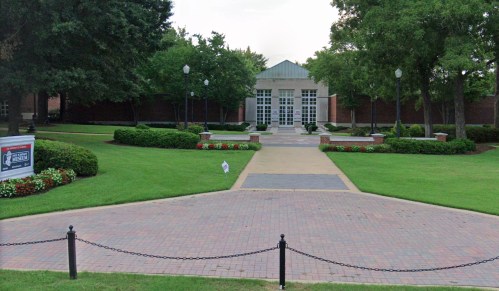 A landscaped courtyard with a brick pathway leading to a modern building, surrounded by trees and flower beds.