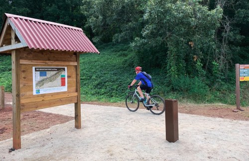 A cyclist rides past a trailhead sign for Red Mountain Trail, surrounded by greenery.