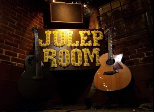 A cozy music venue sign reading "Julep Room" with two acoustic guitars on either side, set against a brick wall.