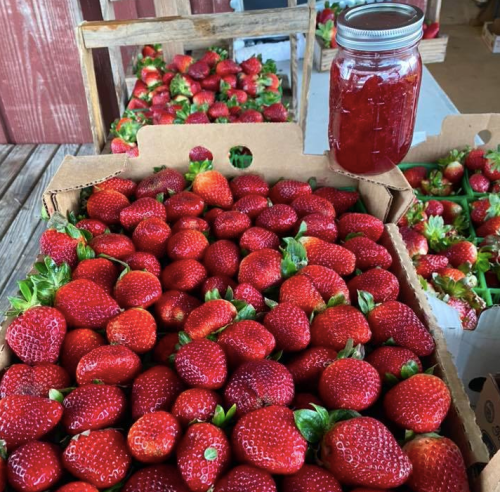 A box of fresh, vibrant red strawberries with a jar of strawberry jam in the background.