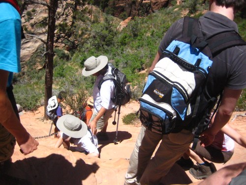 A group of hikers wearing hats and backpacks navigate a rocky trail in a lush, green landscape.